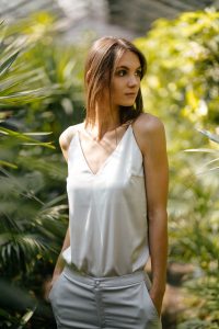 Modelo jovem usando blusa branca ou pérola de seda/cetim com decote em V e alças finas, combinada com calça clara, em um ambiente com folhagens.