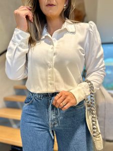 Modelo usando camisa de botão branca de manga longa com detalhes no colarinho e ombros, combinada com calça jeans azul.
